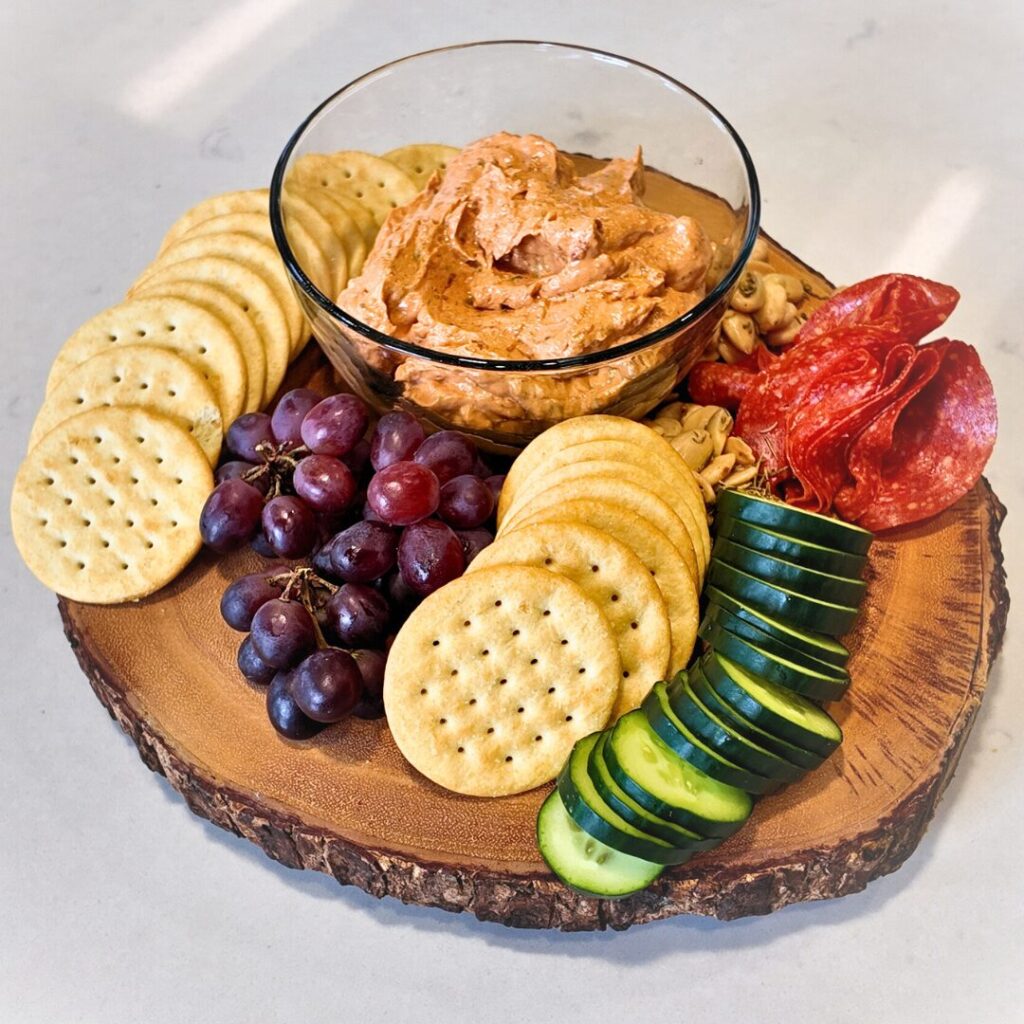 A wooden platter with crackers, sliced cucumber, grapes, salami, nuts, and a bowl of reddish dip in the center.