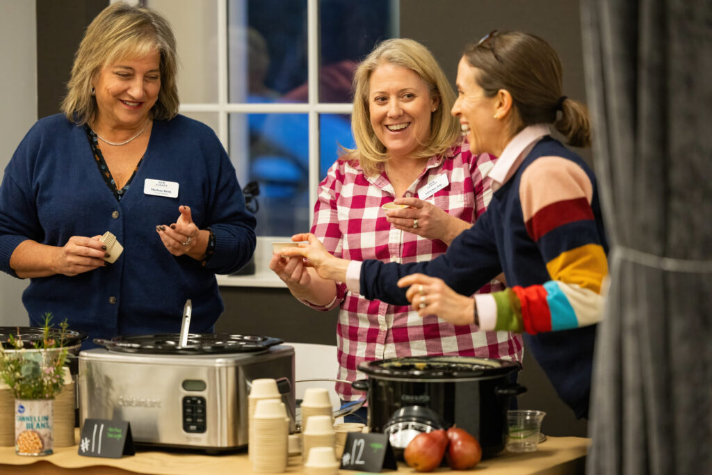 Three women laughing and exchanging small cups of food at a table with crockpots and disposable cups.