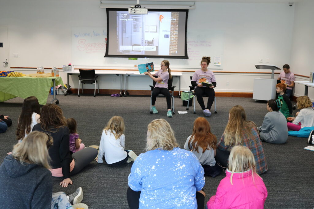 A group of children sit on the floor while two people read books aloud in a room with a projector screen and a table with snacks.