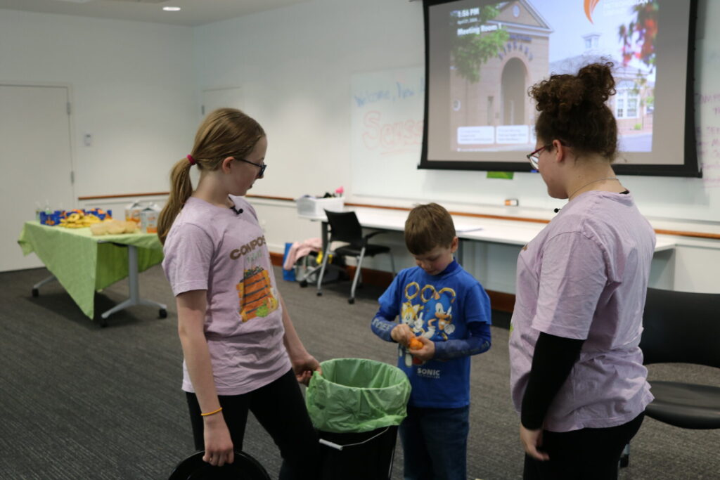 Three children stand in a room with a presentation screen in the background; two are wearing purple shirts and one is peeling an orange.