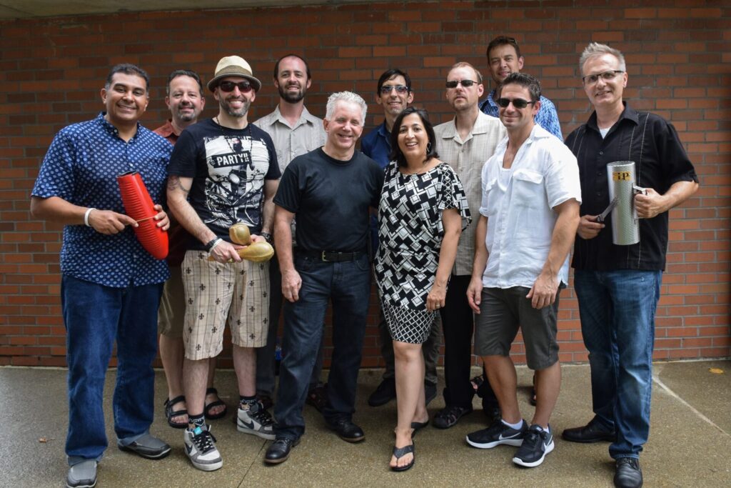 A group of eleven people posing for a photo against a brick wall. Several of them are holding percussion instruments, such as a guiro and maracas. They are casually dressed, and the atmosphere appears friendly and relaxed.