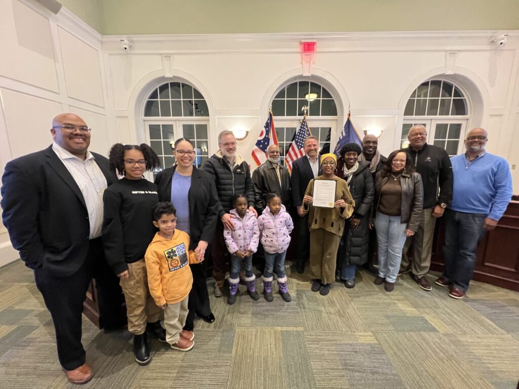 A diverse group of people, including adults and children, posing inside a room with American flags in the background. One person in the center is holding a certificate.