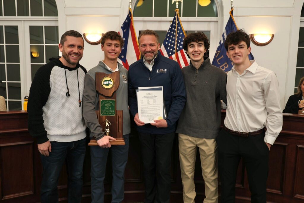 Five people stand together, one holding a trophy labeled "OHSAA 2024 Division II Boys Soccer State Champion," while another holds a certificate. American flags are in the background.