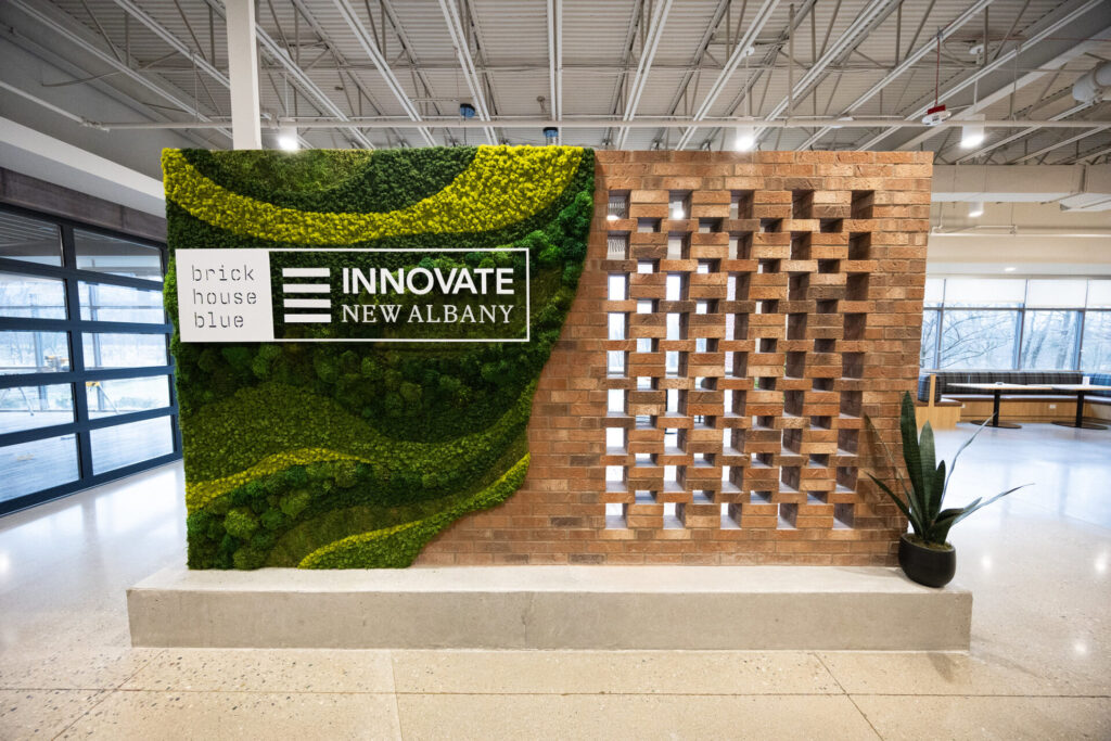 A wall with "brick house blue" and "INNOVATE NEW ALBANY" signage on a green mossy section, adjacent to a perforated brick design with a potted plant at the base.