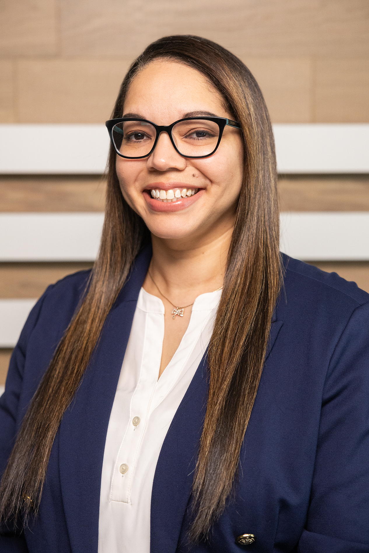 A woman with long straight hair, wearing glasses, a navy blazer, and a white blouse, smiling at the camera in a professional setting.