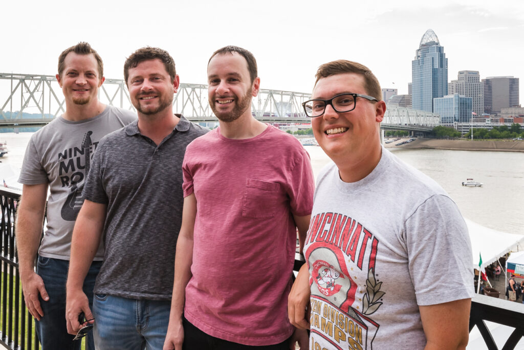 Four men smiling, standing on a waterfront with a bridge and cityscape in the background.