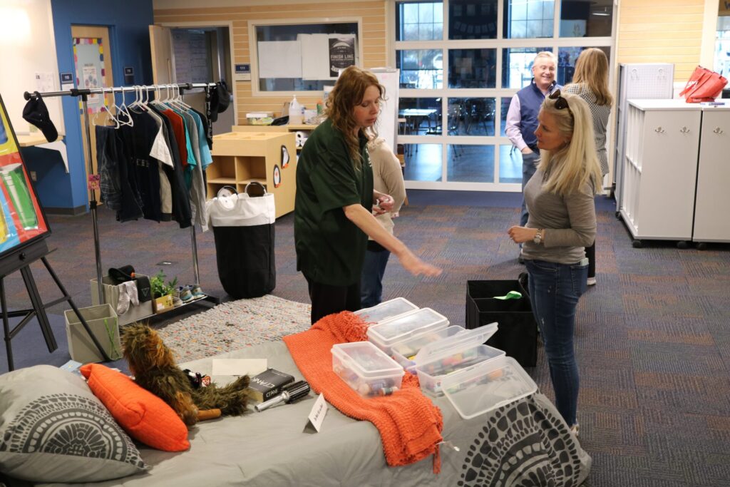 Room with people organizing items; clothes on a rack, storage bins, and household items on a bed.