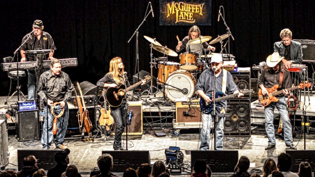 A band performs on stage with a variety of instruments, including guitars, drums, and keyboard, under a sign that reads 'McGuffy Lane.'