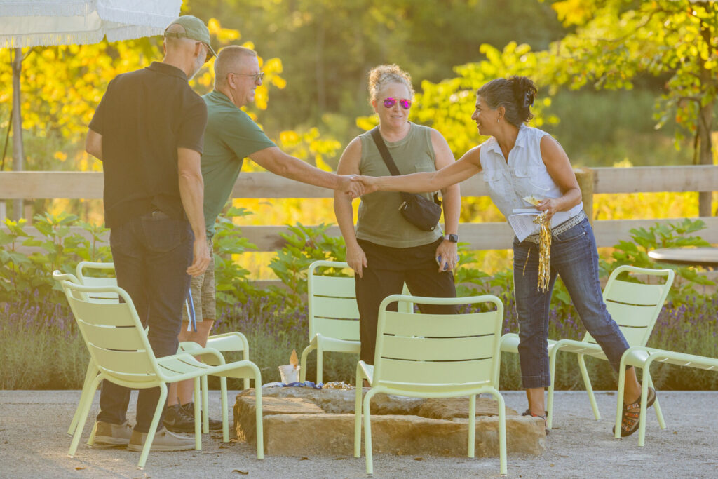 Four people standing outdoors, one shaking hands with another, amidst green chairs and a wooden fence in the background on a sunny day.