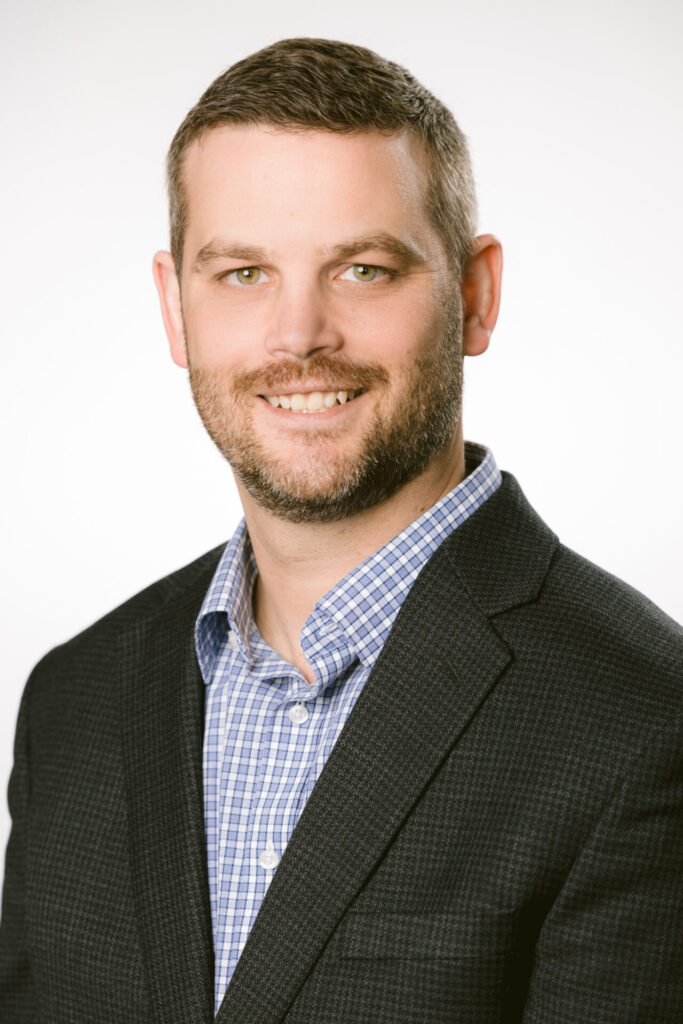 Smiling man with short hair and beard, wearing a dark blazer and blue checked shirt against a white background.