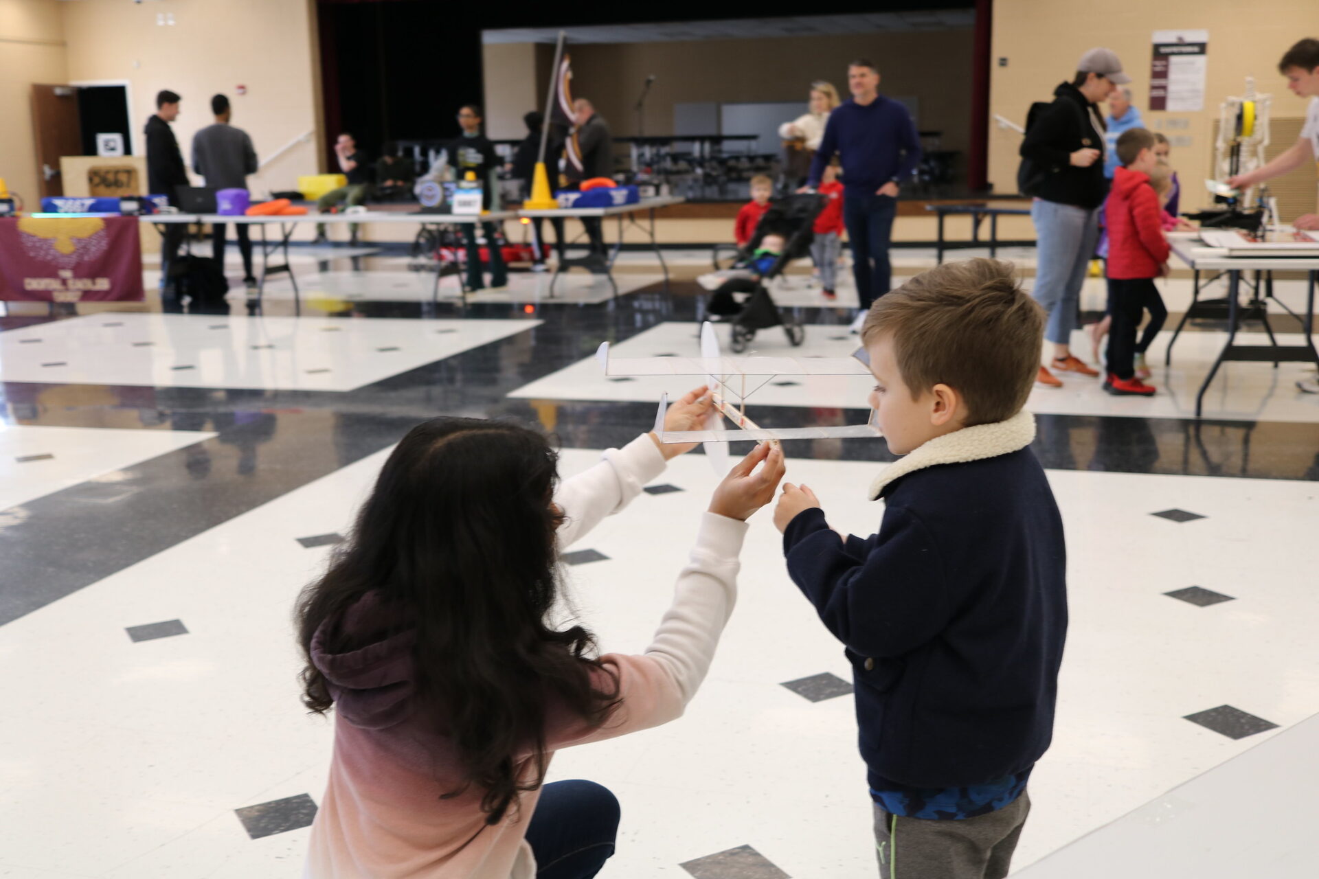 A woman kneels to show a young boy a small model airplane in a large indoor hall with various tables and people in the background.