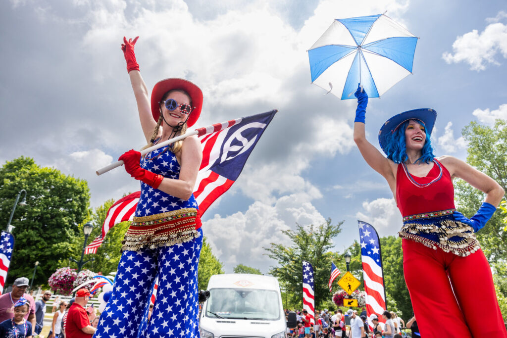 Two women dressed in patriotic red, white, and blue costumes with star patterns, standing on stilts at a festival. One holds an American flag, and the other holds a blue and white umbrella. People and American-themed decorations are visible in the background under a partly cloudy sky.