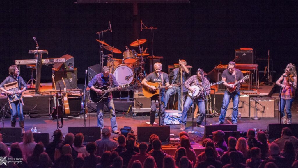 A band performs on stage with various instruments including guitars, a banjo, a violin, and a washboard, in front of an audience.