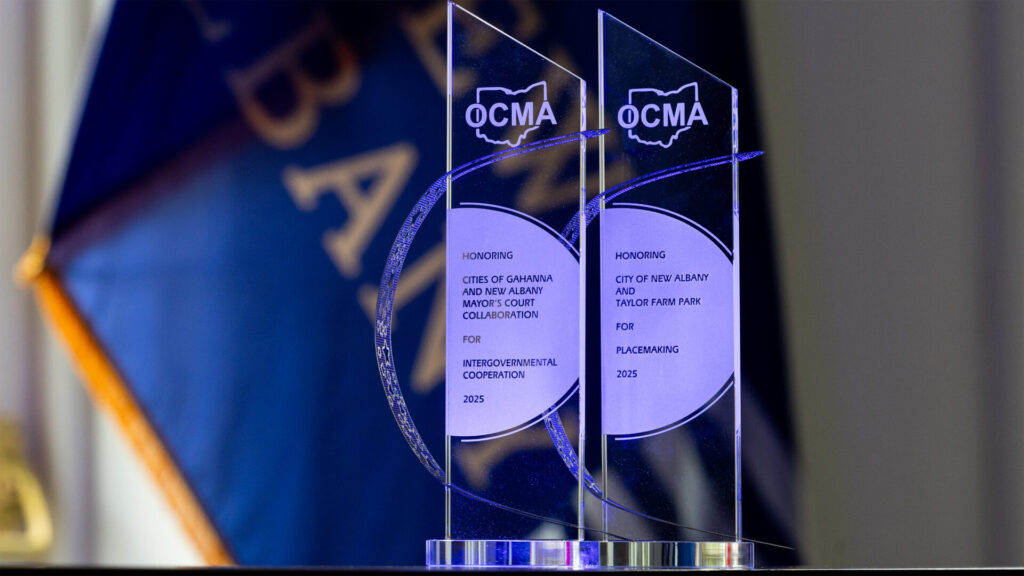 Two clear award plaques displaying the OCMA logo, honoring cities for intergovernmental cooperation and placemaking in 2025, with a blurred flag in the background.