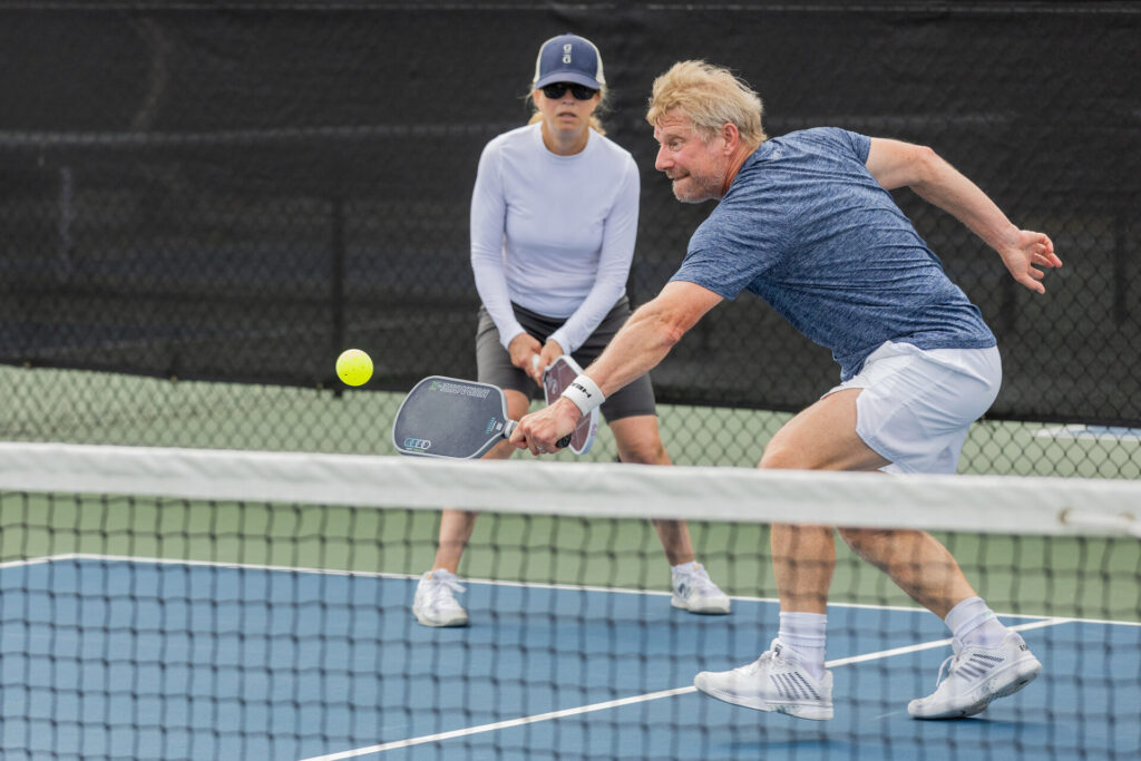 Two people playing pickleball on an outdoor court, with one player hitting the ball towards the net while the other watches.