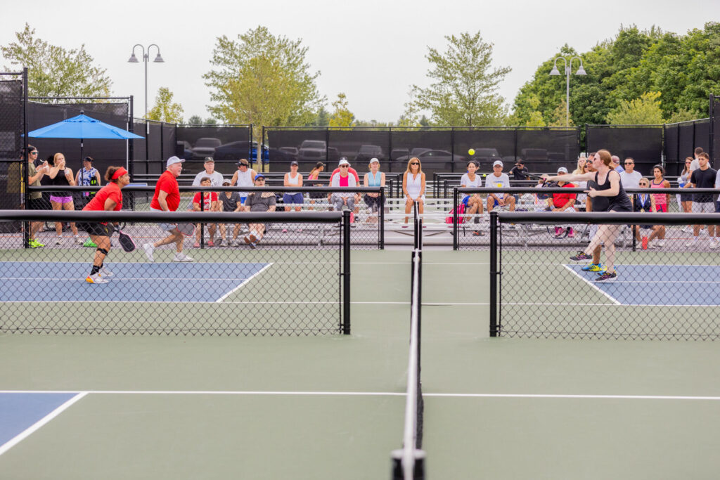 A pickleball game in progress with two players in red shirts on the left and two players in black outfits on the right, while a group of spectators sits in the background on bleachers under a cloudy sky.