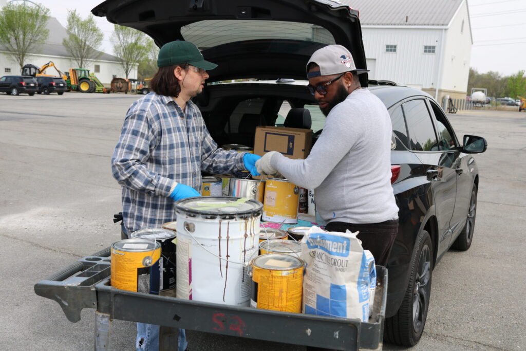 Two individuals next to a car loading paint cans and supplies onto a cart, with a background of a parking lot and agricultural equipment.