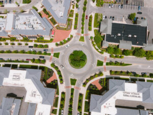 Aerial view of a roundabout surrounded by buildings and greenery, with multiple cars navigating the circular intersection.