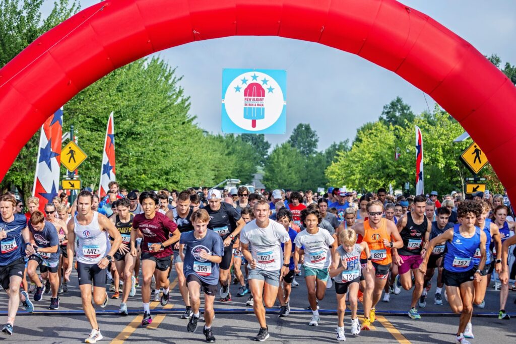 A large group of runners starting a race on a tree-lined street under a red inflatable arch with a sign that reads New Albany 5K Run & Walk, with participants of various ages wearing numbered bibs and athletic clothing.