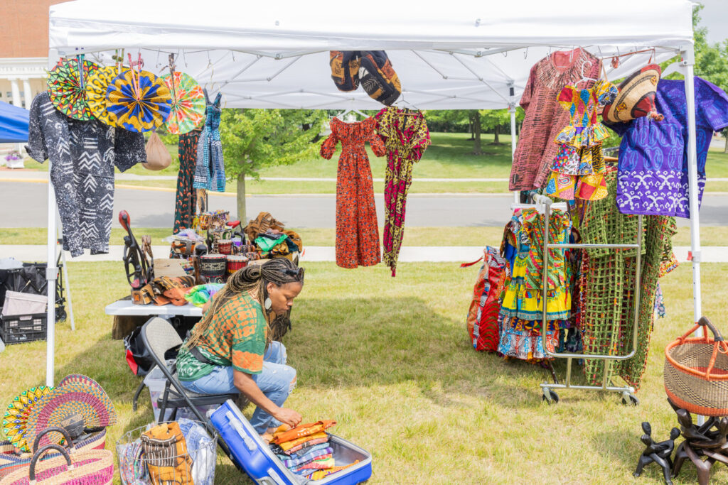 A woman organizing colorful clothing and accessories at an outdoor market stall under a white canopy.