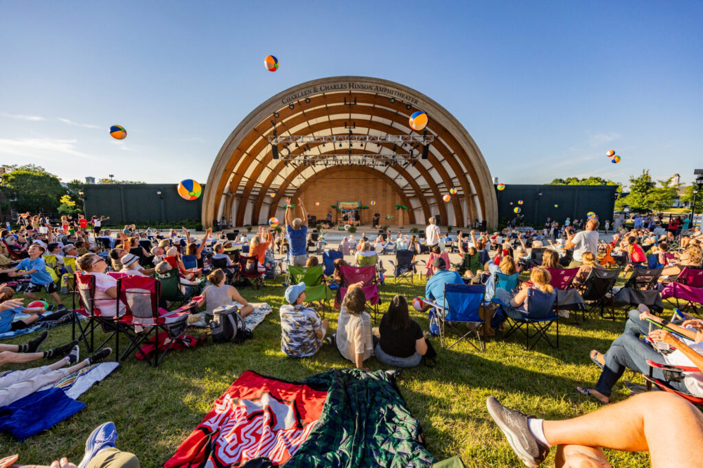 A lively outdoor concert at an amphitheater with a large audience seated on the grass, some on folding chairs. Beach balls are being tossed around, adding to the festive atmosphere. The stage is set against a clear blue sky, suggesting a sunny day.