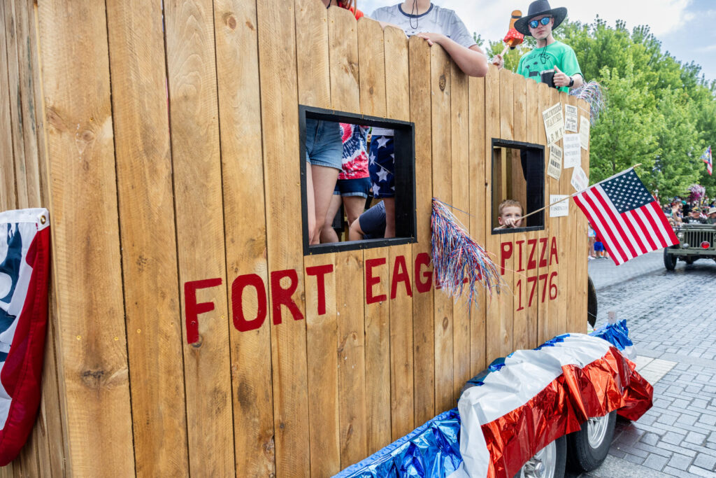 Parade float designed as a wooden fort with people inside and on top, decorated with red, white, and blue streamers and American flags, featuring the text "FORT EAGLE PIZZA 1776" painted in red on the side.