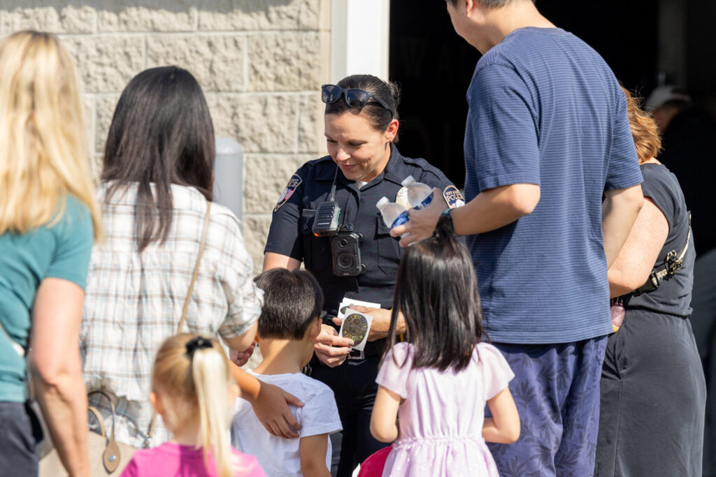 A police officer interacting with a group of children and adults outdoors, handing out a sticker to a child in a white shirt.