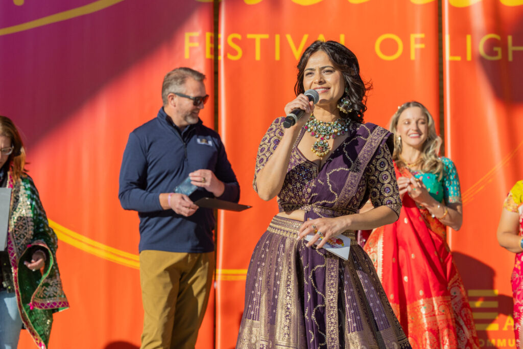 A woman in a purple traditional dress is speaking into a microphone at a festival. She is surrounded by people in colorful traditional attire. The backdrop reads "Festival of Light."