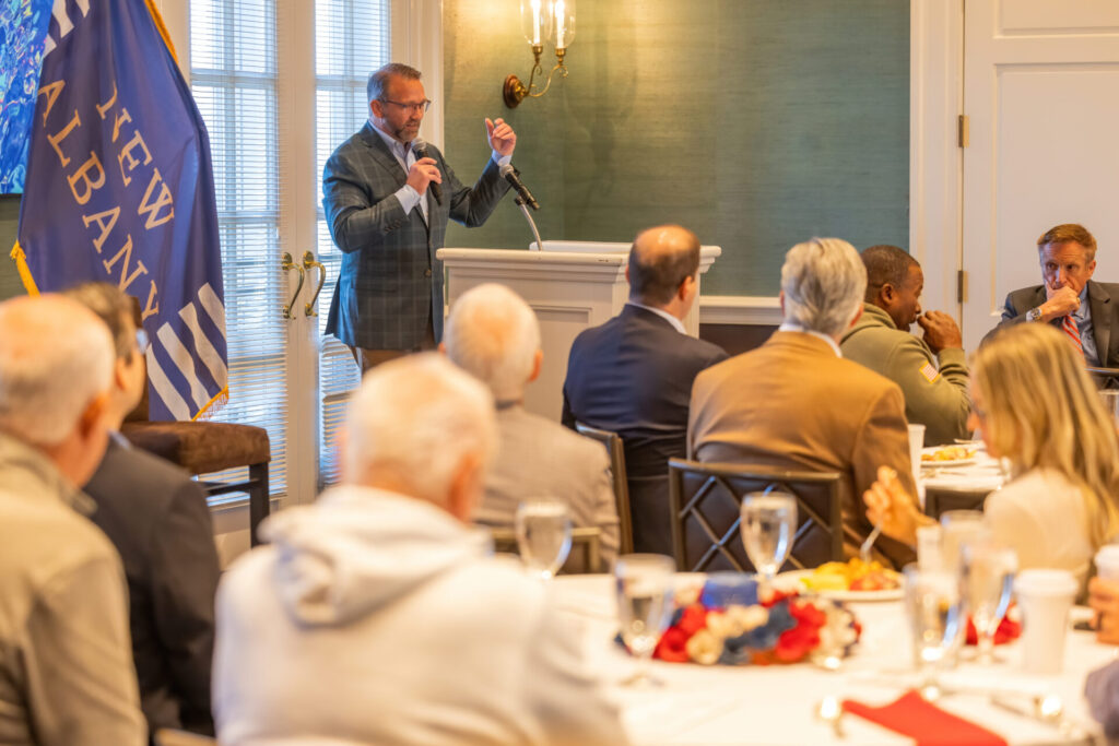 A man speaking at a podium in a formal setting, with an audience seated at tables in front of him. The room features a New Albany flag and decorative lighting, suggesting a community or business event.