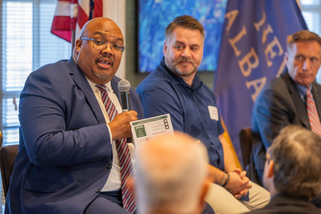 A man in a blue suit holds a microphone and speaks at an event, sitting beside two other men; one in a blue shirt and another in a grey suit. A flag is displayed in the background.