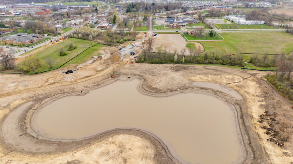 Aerial view of a large construction site with a newly formed pond surrounded by dirt and earthmoving activity, green fields, houses, and buildings in the background under a cloudy sky.