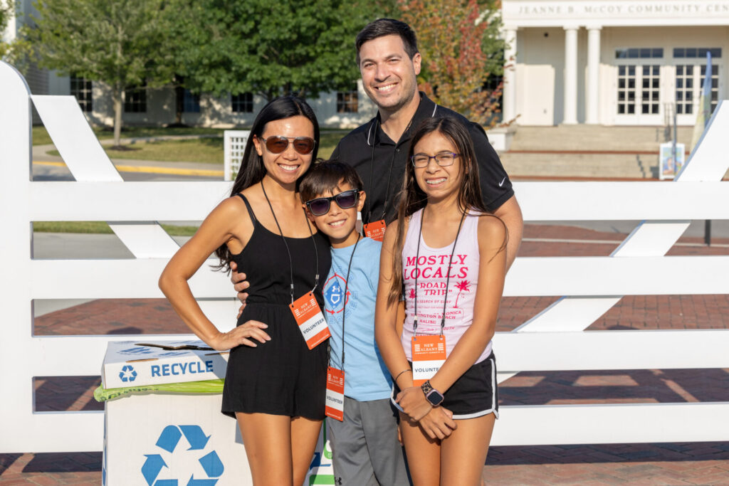 A family of four, consisting of two adults and two children, all wearing sunglasses and volunteer badges, pose smiling in front of a building labeled "Jeanne B. McCoy Community Center." They stand near a recycling bin and a white fence, with trees in the background.