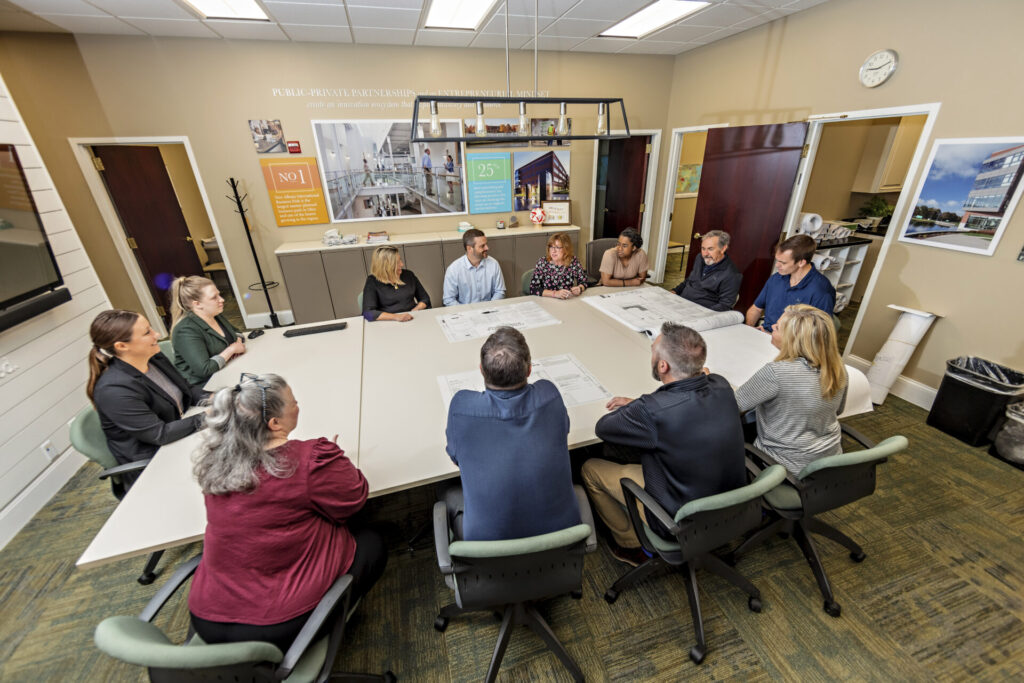 Eleven people seated around a large conference table in a meeting room, discussing architectural blueprints spread out on the table, with office doors, wall art, and a clock visible in the background.