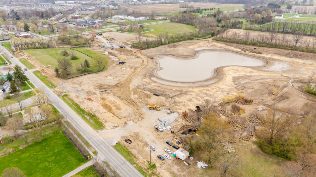Aerial view of a construction site with a large, irregularly shaped water retention pond surrounded by dirt and construction equipment, adjacent to a road and fields with scattered trees and residential buildings in the background.