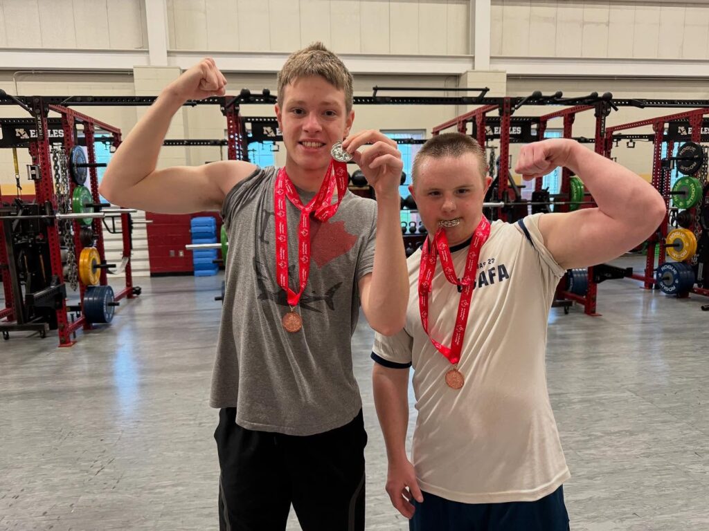 Two young men in a gym flexing their arm muscles and proudly displaying multiple red-labeled medals around their necks, standing in front of weightlifting equipment racks.