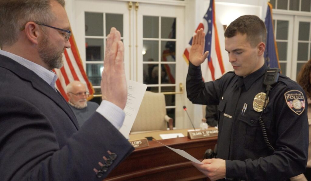 A police officer raising his right hand and taking an oath administered by a man also raising his right hand, inside a formal room with American flags in the background and a man sitting behind a desk.
