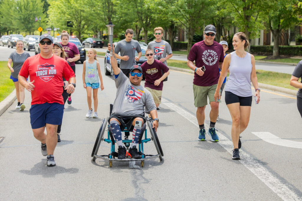 A group of people participating in an outdoor torch run event, including a person in a wheelchair holding a torch, others running and walking on a tree-lined street, some wearing event T-shirts and athletic wear.