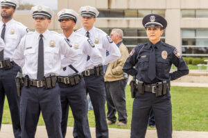 Group of uniformed police officers, including one female officer in dark uniform and multiple male officers in white shirts and dark ties, standing outdoors in a formal stance with hands behind their backs near a building with glass windows and green grass.