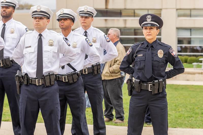Group of uniformed police officers, including one female officer in dark uniform and multiple male officers in white shirts and dark ties, standing outdoors in a formal stance with hands behind their backs near a building with glass windows and green grass.