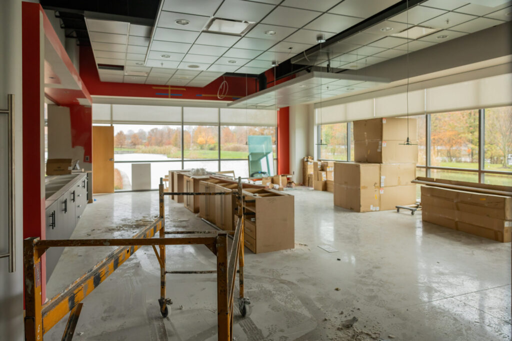 Interior of a room under construction with large windows overlooking a pond and autumn trees, featuring unpacked cabinets and stacked cardboard boxes on a dusty floor with construction barriers in the foreground.
