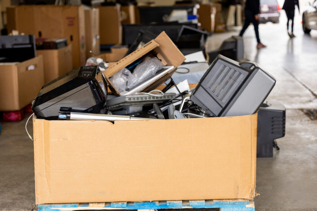 Large cardboard box filled with discarded electronic devices including printers, a CRT monitor, and other computer peripherals in a warehouse setting with blurred people and boxes in the background.