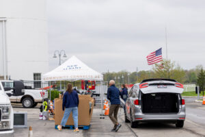 People setting up or organizing items under a white tent labeled 'New Albany Community Connects Us' next to a silver minivan with its rear hatch open, with an American flag flying in the background on a cloudy day.