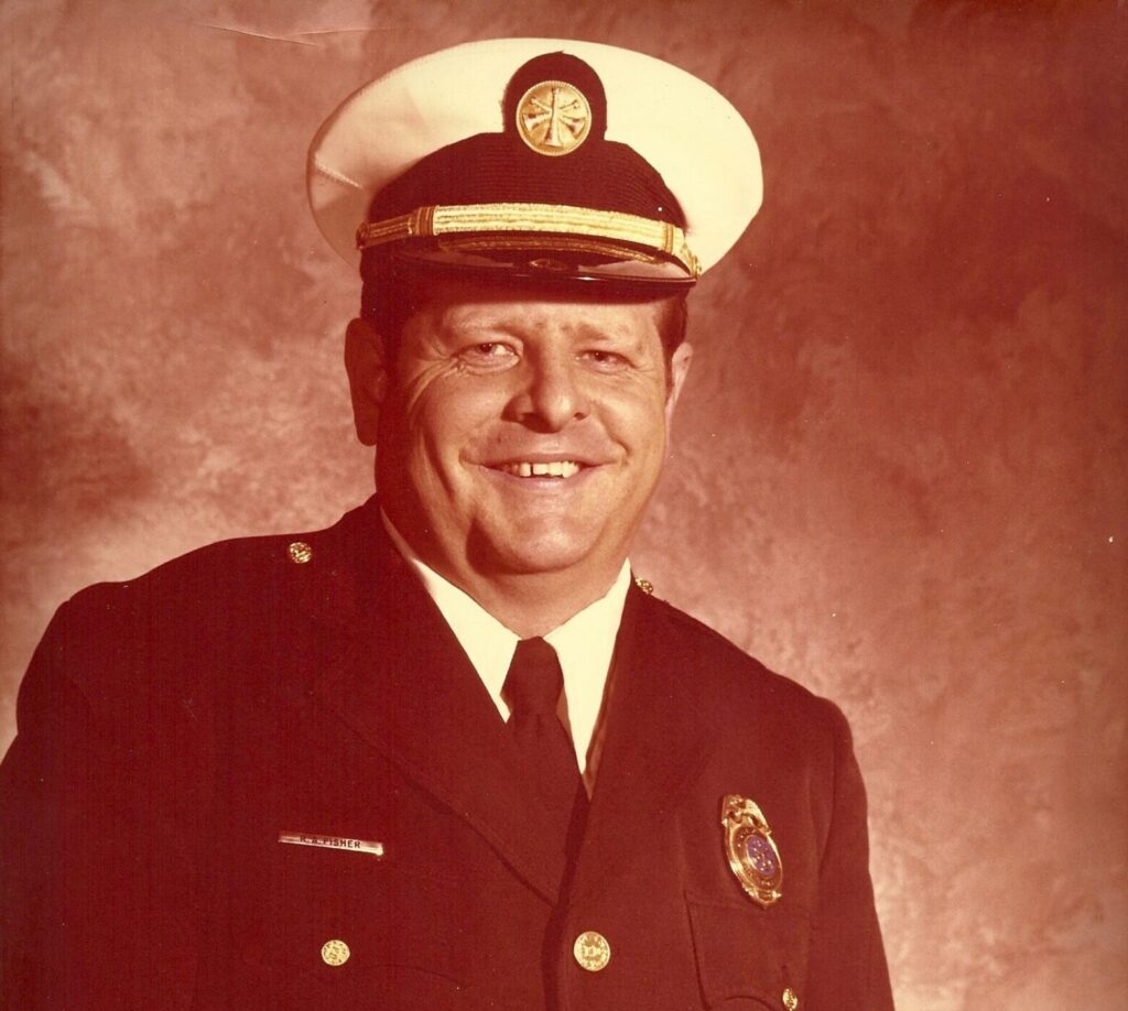 Smiling man in a dark formal uniform with gold buttons and a badge, wearing a white ceremonial hat with a gold emblem and a black visor, standing against a textured brown background.