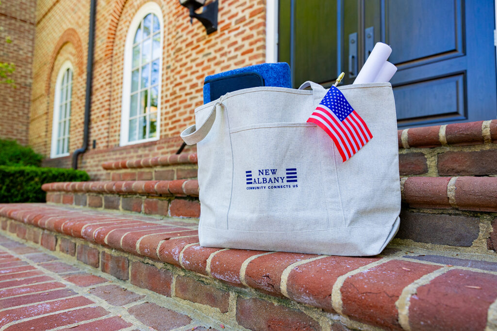 A beige tote bag with the text New Albany Community Connects Us sits on brick steps outside a building, holding an American flag, rolled papers, and a blue item inside.