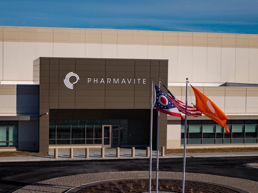 Exterior view of a modern building with the logo and name PHARMAVITE on a dark brown facade, accompanied by three flagpoles flying the Ohio state flag, American flag, and an orange PHARMAVITE flag, under a clear blue sky.