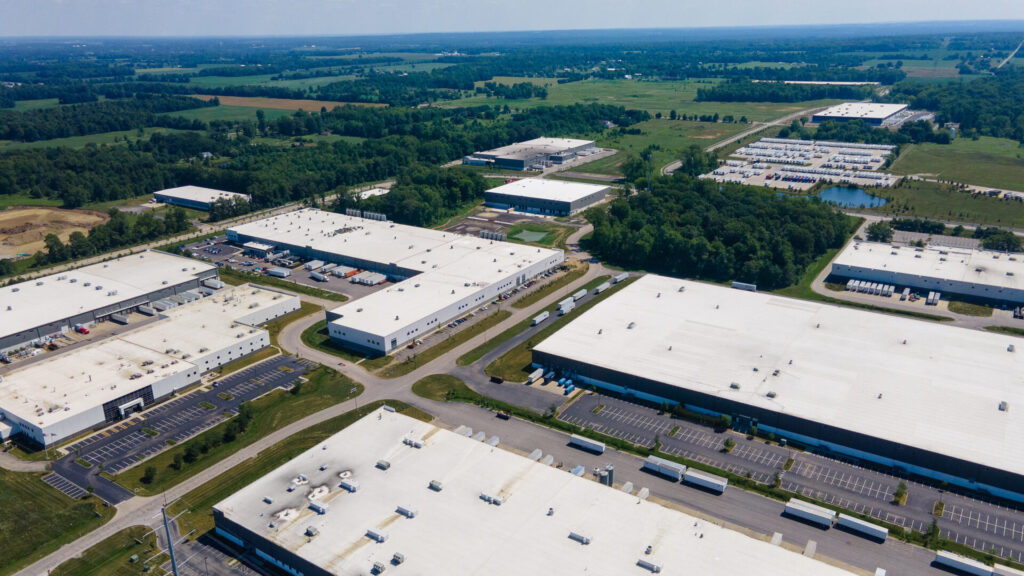 Aerial view of a large industrial park with multiple warehouses featuring white roofs, surrounded by parking lots, roads, and green fields extending into the distance under a clear sky.