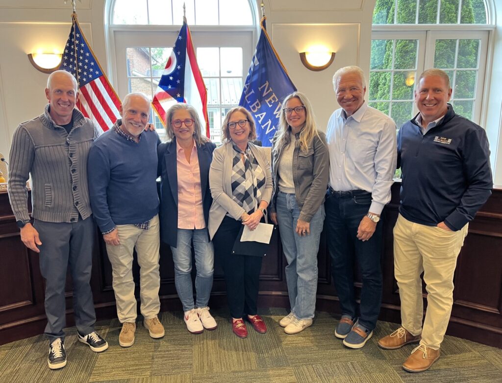 Seven adults standing indoors side by side in front of three flags, including the United States flag and Ohio state flag, with windows in the background. They are casually dressed and smiling at the camera.