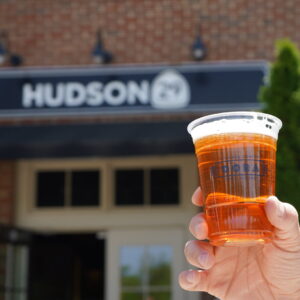 Hand holding a plastic cup filled with amber beer in front of a brick building with a sign that reads HUDSON.