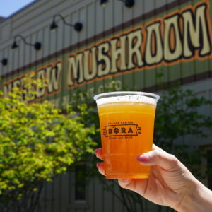 Hand holding a plastic cup of orange beer with foam, in front of a building with a sign reading 'Old New Mushroom' and greenery around it