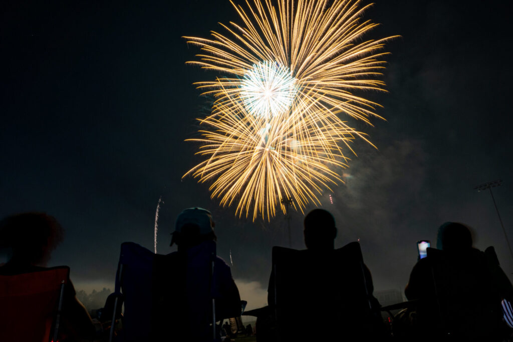 Silhouetted people sitting in chairs watching golden and white fireworks exploding in the night sky.